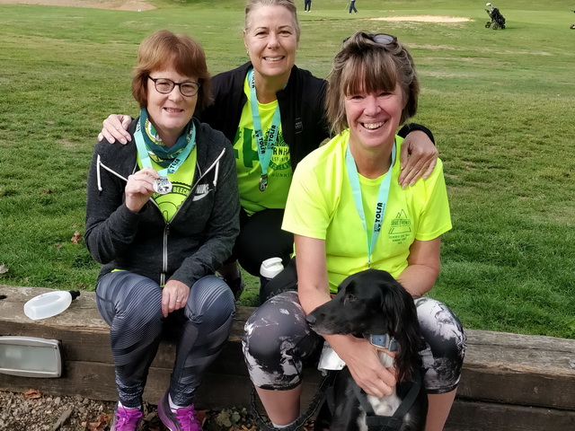 Three women in running gear sit in a park smiling for the camera